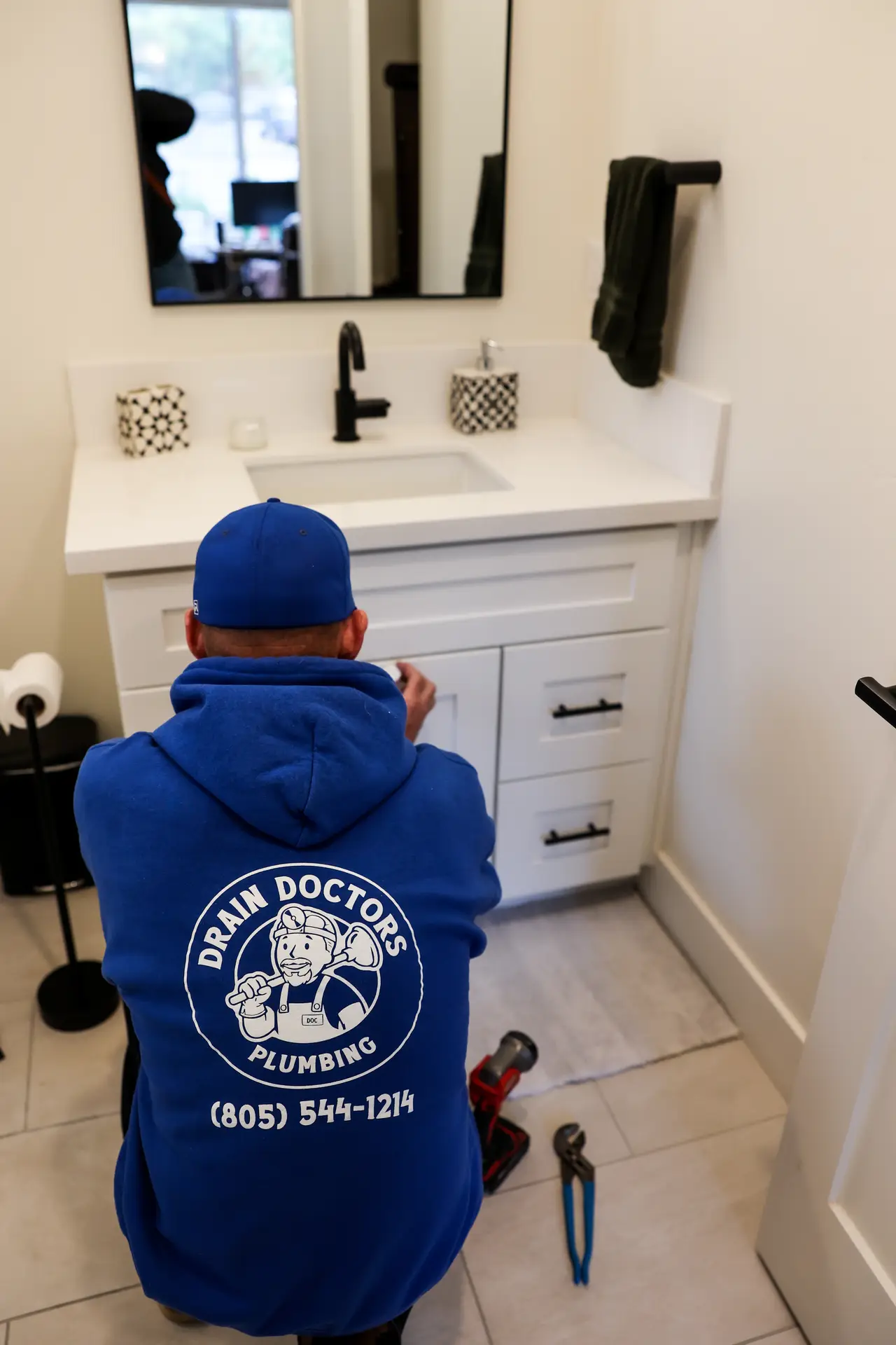 Drain Doctors Plumbing technician kneeling in front of a bathroom vanity with tools on the floor