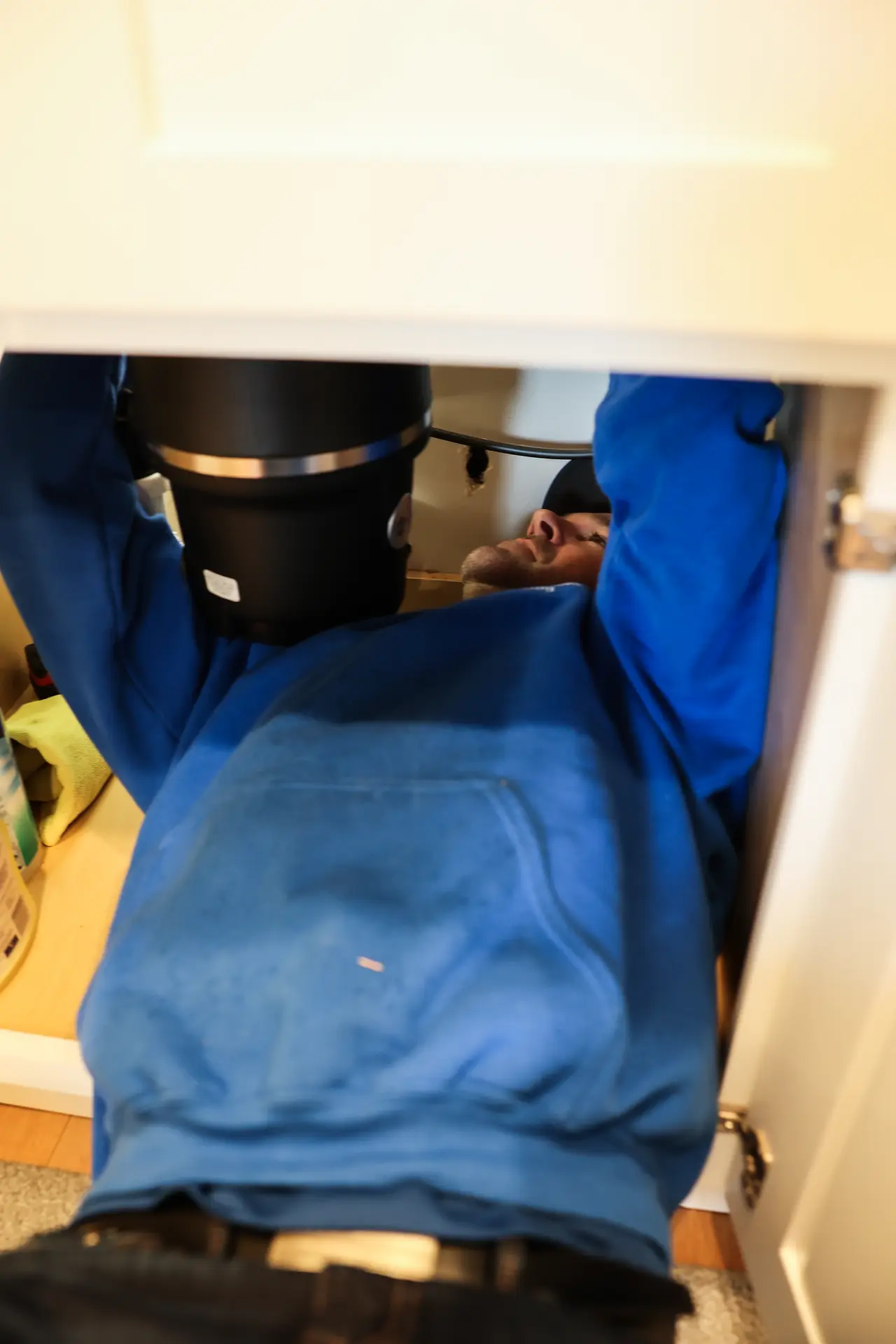Plumber lying inside a kitchen sink cabinet reaching up to work on a black garbage disposal