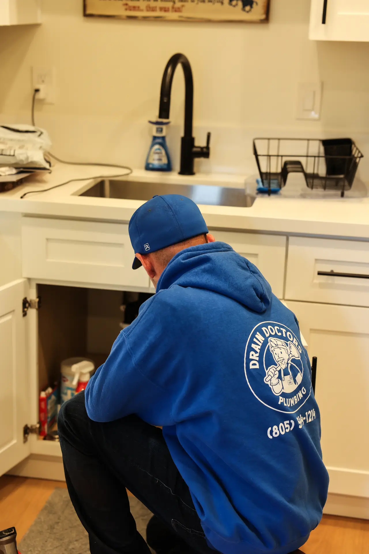 Drain Doctors Plumbing technician kneeling at an open kitchen sink cabinet to inspect the plumbing underneath