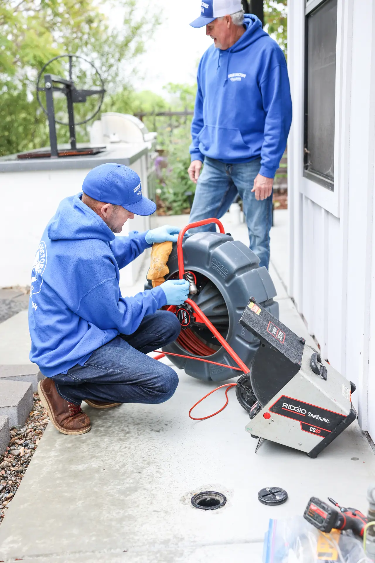 Two Drain Doctors Plumbing technicians setting up a sewer camera reel next to an open cleanout beside a home