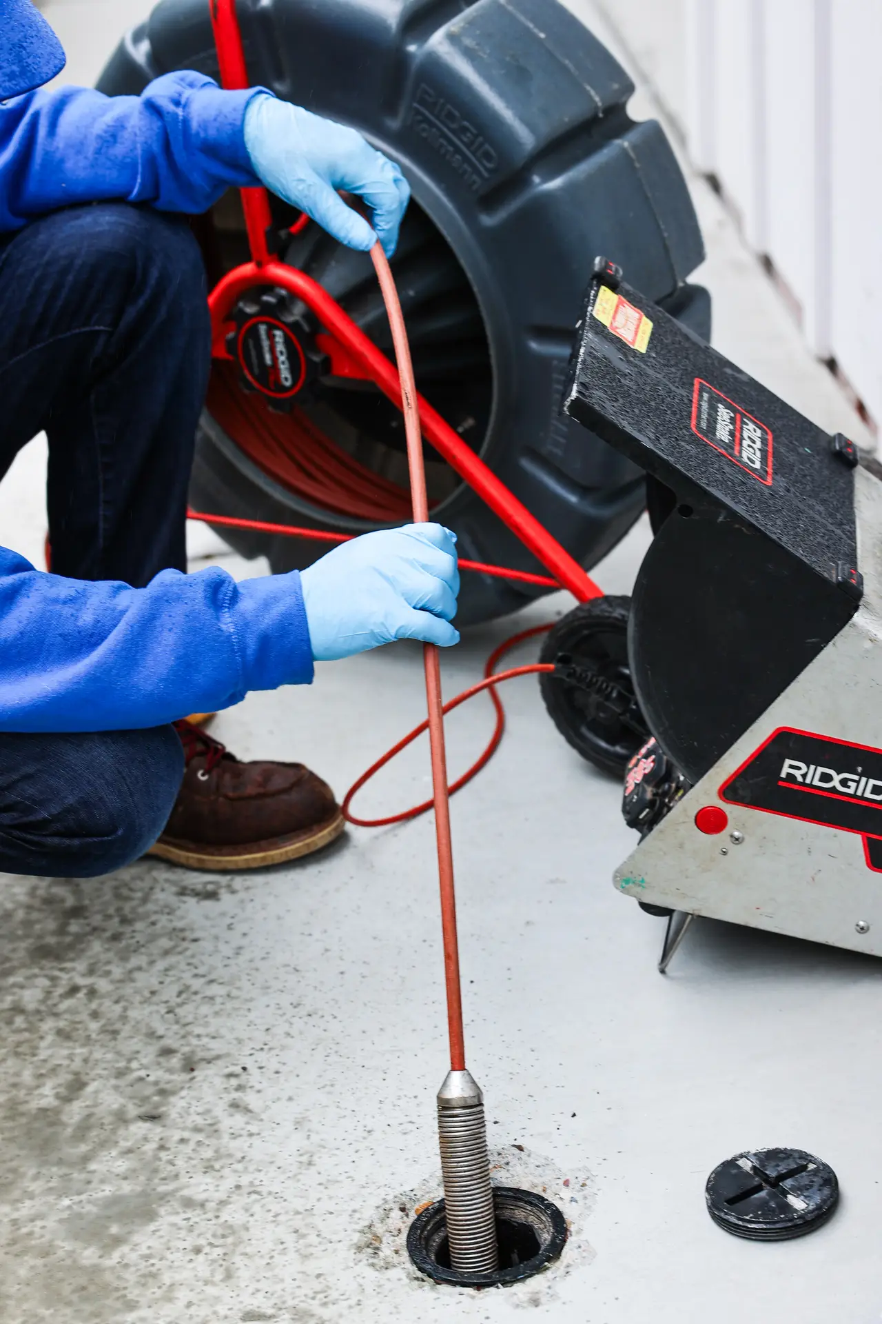 Close-up of a technician's gloved hands guiding a sewer camera cable from the reel into an outdoor cleanout