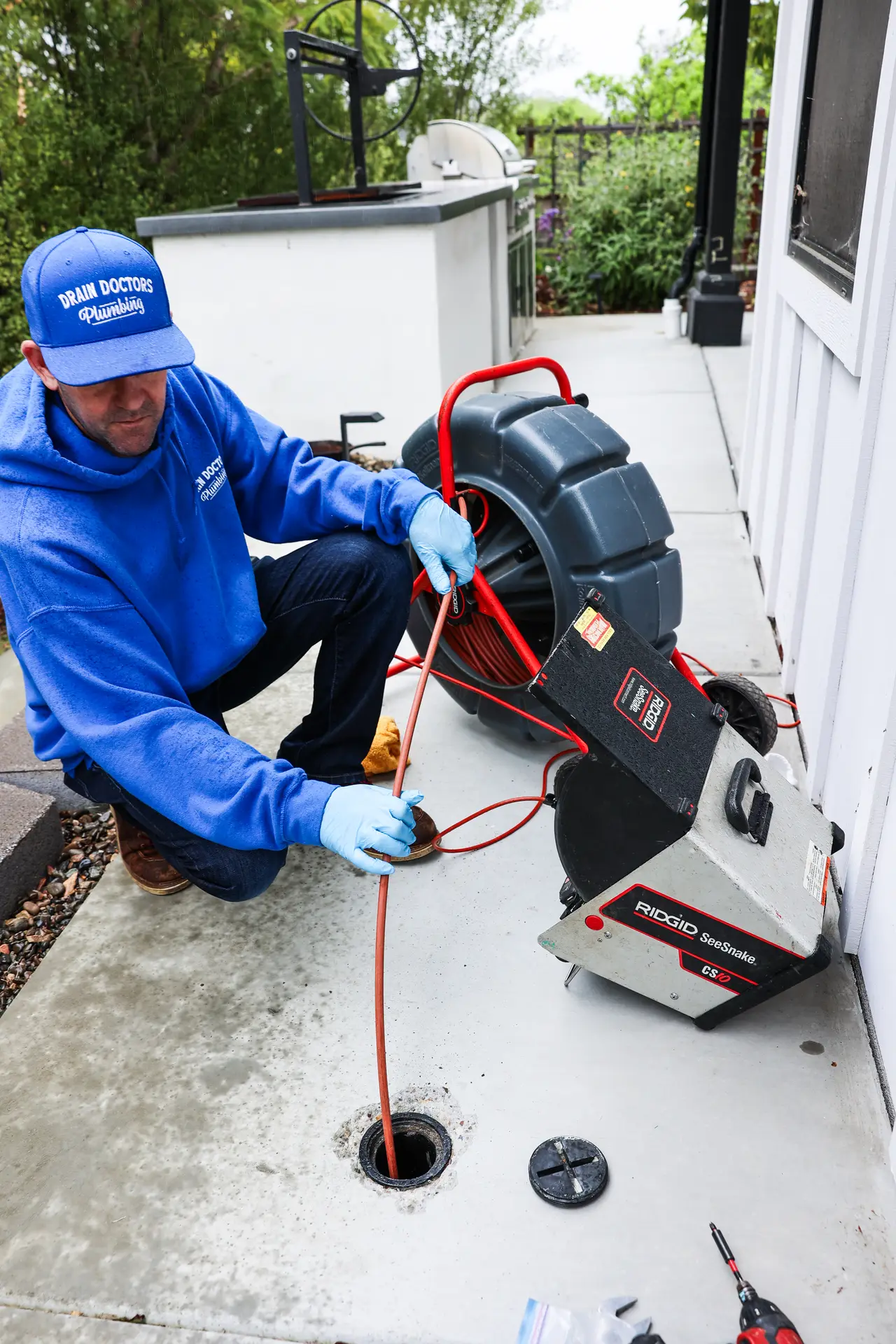 Drain Doctors Plumbing technician kneeling beside a sewer camera reel and feeding the cable into a backyard cleanout