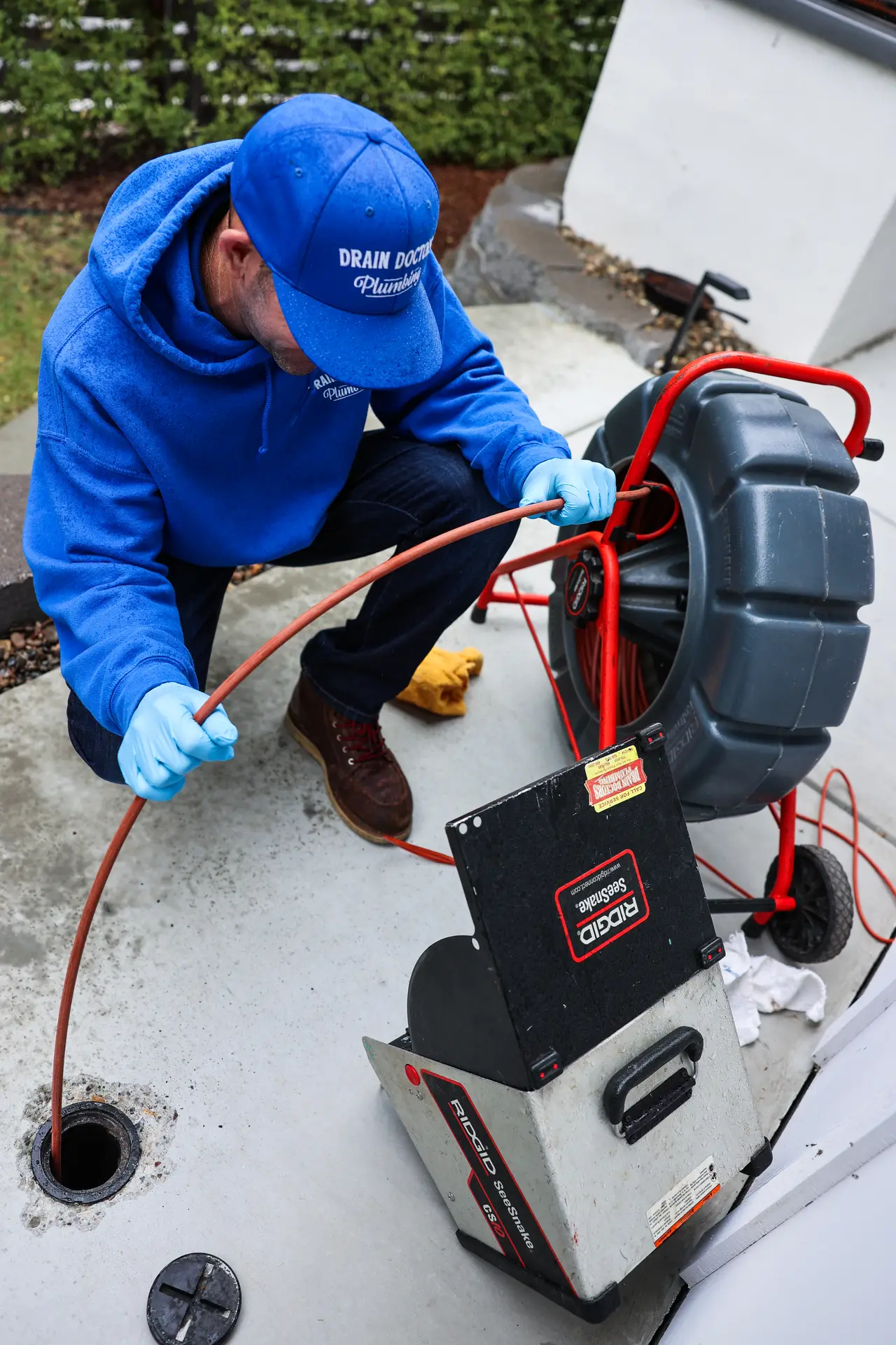 Drain Doctors Plumbing technician leaning over a camera reel and guiding the inspection cable into a sewer cleanout