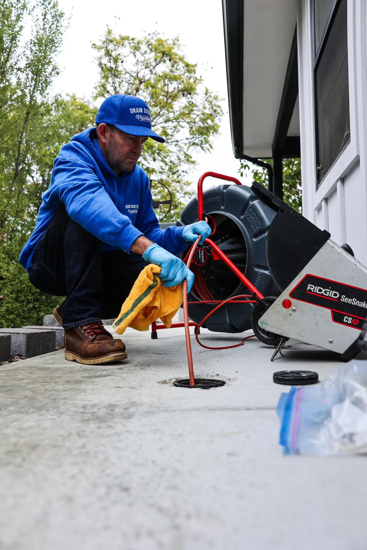 Drain Doctors Plumbing technician feeding a sewer camera cable into a cleanout beside a home, with mature trees in the background