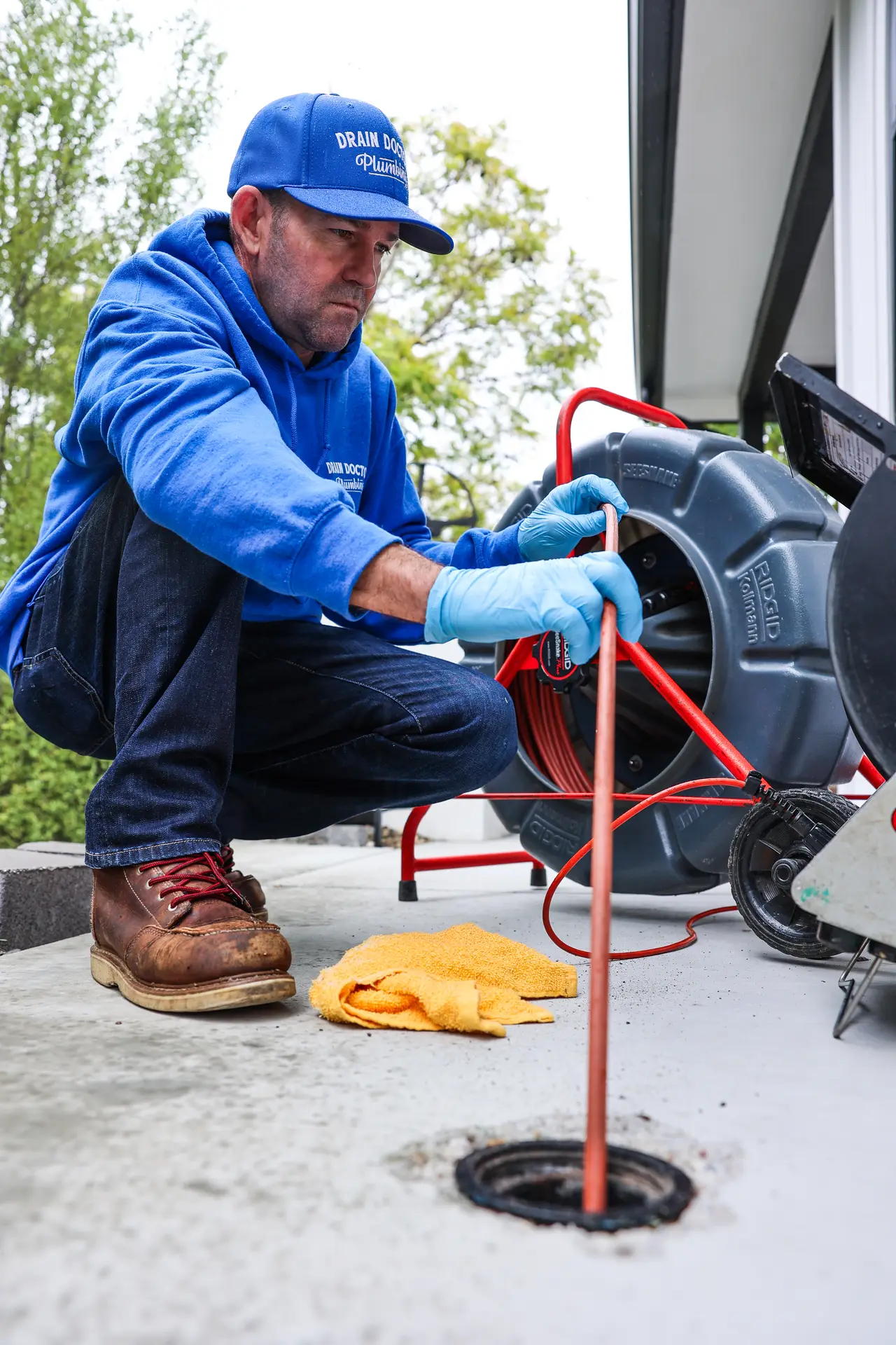 Drain Doctors Plumbing technician kneeling beside a camera reel and feeding the inspection cable into a sewer cleanout