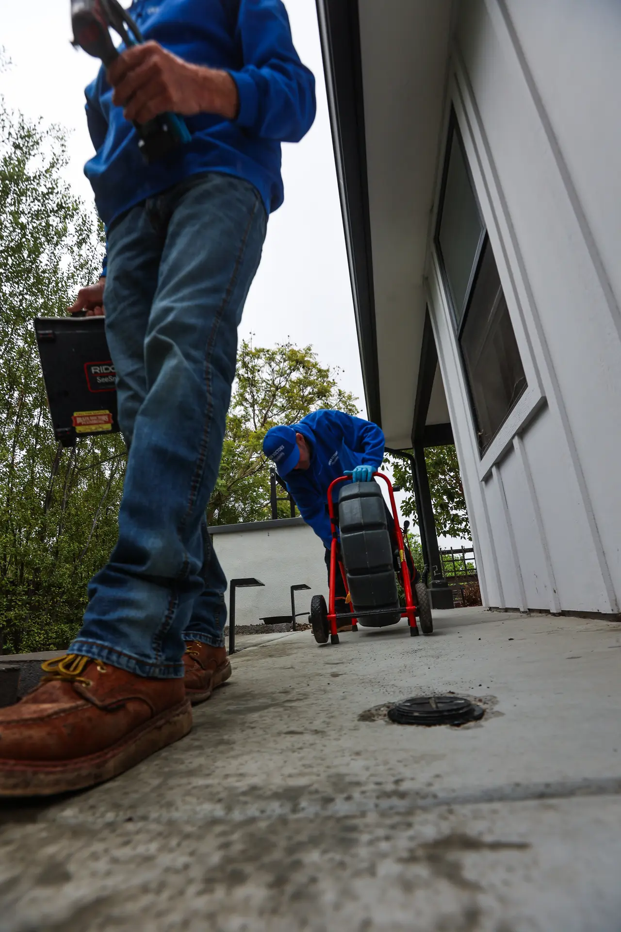 Two Drain Doctors Plumbing technicians arriving at an outdoor cleanout with a sewer camera reel and monitor