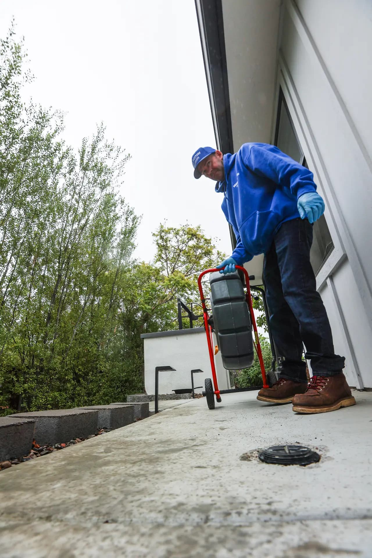 Drain Doctors Plumbing technician wheeling a sewer camera reel toward an open cleanout alongside a house