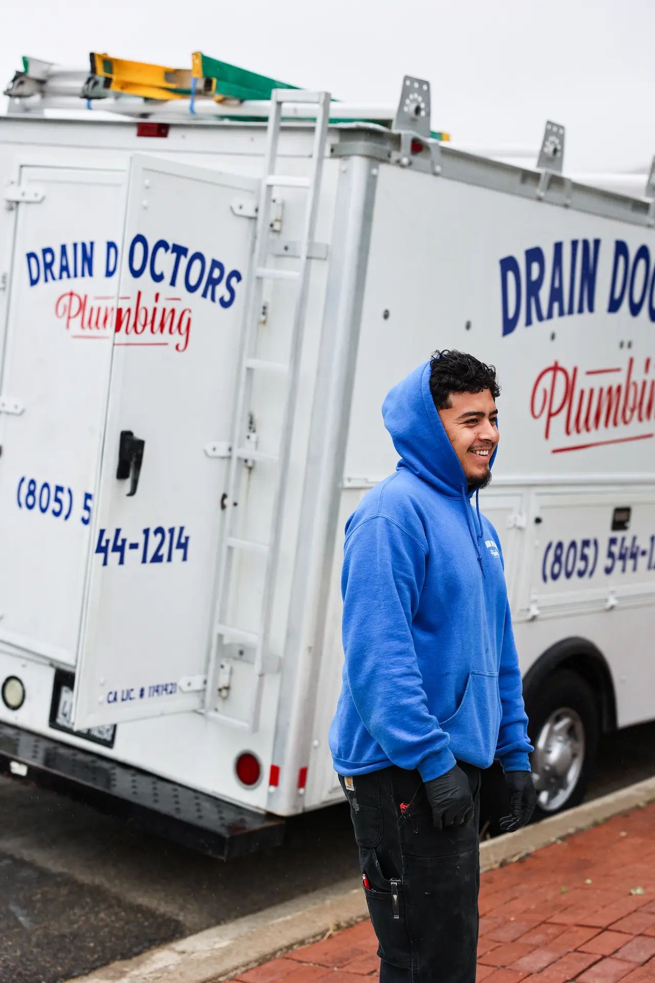 Smiling Drain Doctors Plumbing technician in a blue hoodie and work gloves standing beside the service truck