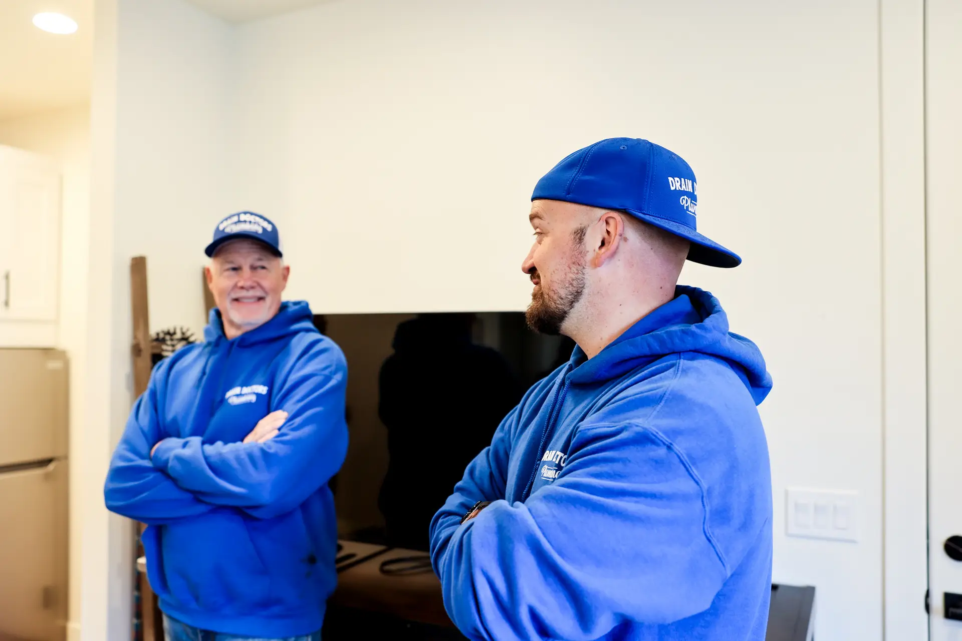 Two Drain Doctors Plumbing technicians in blue hoodies talking inside a customer's home near the kitchen