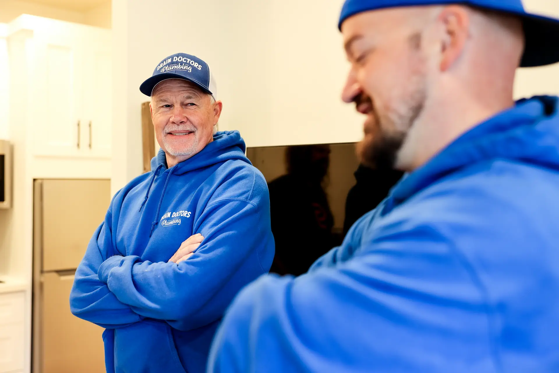 Smiling Drain Doctors Plumbing technician with arms crossed standing in a customer's kitchen next to a coworker