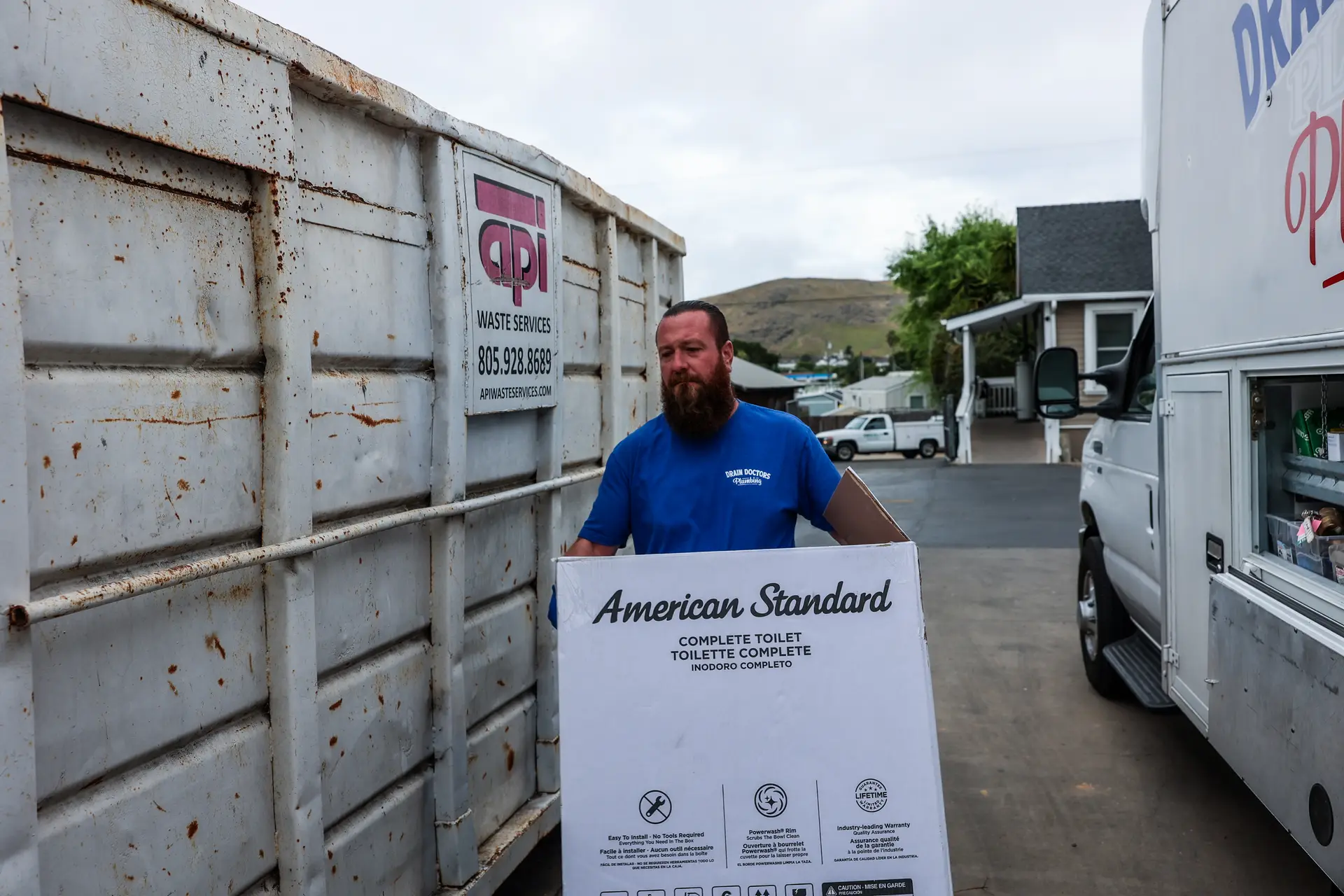 Drain Doctors Plumbing technician carrying a boxed American Standard toilet past the service truck