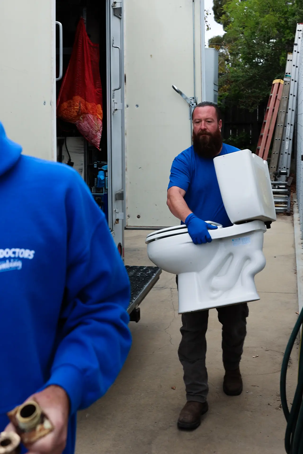 Drain Doctors Plumbing technician in blue gloves carrying a removed toilet past the open door of a service truck